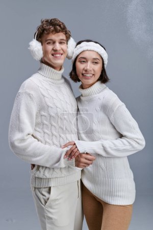 smiley interracial couple in cozy knitted sweaters posing on grey backdrop with snow, trendy winter