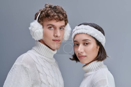 fashion lookbook concept, portrait of interracial couple in winter outfit looking at camera on grey