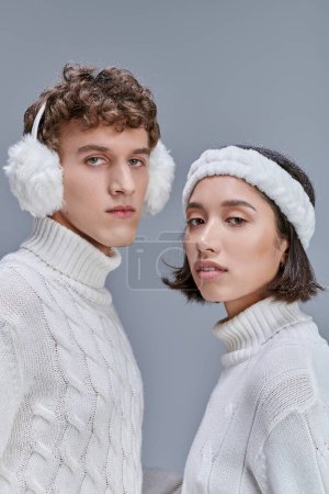 winter fashion concept, portrait of multiethnic couple in warm outfit looking at camera on grey