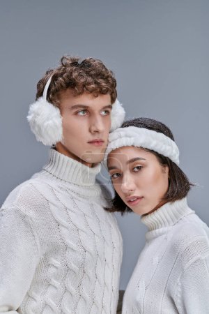 joven asiático mujer con nevado pelo mirando a cámara cerca hombre en caliente orejeras en gris, invierno estilo
