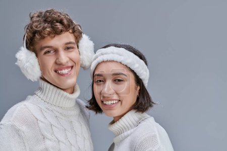 cheerful interracial couple in winter sweaters posing with snowy hair and looking at camera on grey