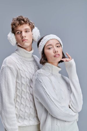 young and stylish multiethnic couple in white cozy sweaters posing with snowy hair on grey backdrop