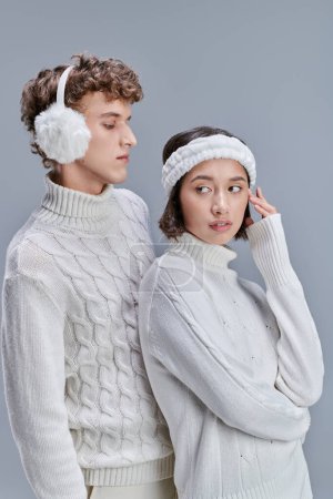 mujer asiática elegante mirando lejos cerca de hombre joven en orejeras calientes sobre fondo gris, pelo cubierto de nieve