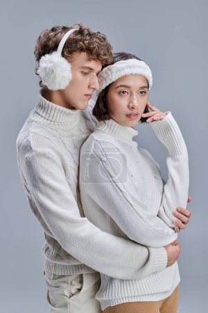 junger Mann mit Ohrenschützer umarmt charmante asiatische Frau auf grauem Hintergrund, romantisches Paar im Winter