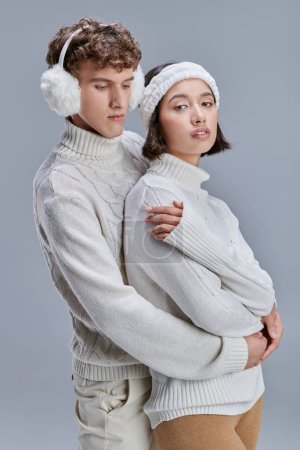 trendy man in earmuffs hugging young asian woman on grey background, stylish romantic winter