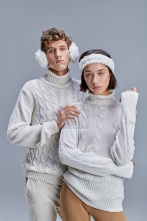 fashionable dressed interracial couple looking at camera on grey backdrop, winter fashion concept