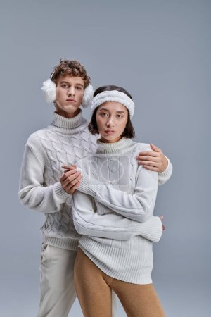 Mann mit Winterohrenschützer umarmt trendige asiatische Frau im Pullover auf grauem, modischem Lookbook-Konzept
