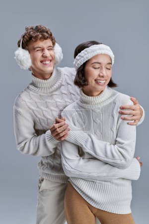 overjoyed man in cozy earmuffs hugging asian woman in winter sweater on grey, seasonal style