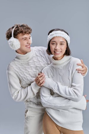 cheerful man in warm earmuffs embracing asian woman in winter sweater on grey, seasonal fashion