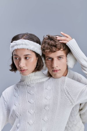 asiatische Frau berührt schneebedecktes Haar eines Mannes in warmen Ohrenschützern vor grauem Hintergrund, romantischer Winterstil