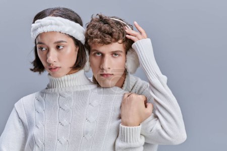 charming woman touching snowy hair of man in warm earmuffs on grey background, coxy winter style
