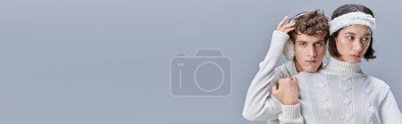homme à la mode en cache-oreilles regardant la caméra près de femme asiatique toucher ses cheveux neigeux sur gris, bannière