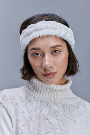 portrait of young asian woman in warm headband and white knitted sweater looking at camera on grey