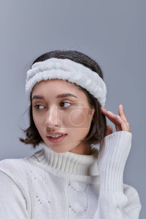 stylish asian woman in warm headband and white sweater posing on grey, winter fashion concept