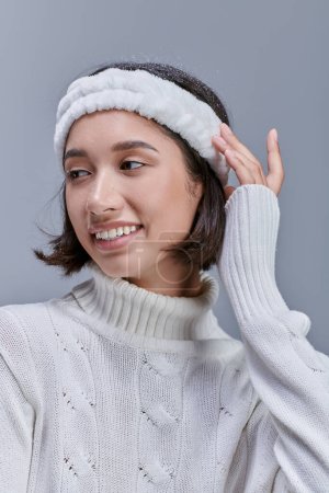 winter fashion campaign, cheerful asian model in knitted sweater and headband looking away on grey