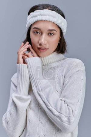 sensual asian woman with snowy hair and warm headband looking at camera on grey, urban winter style