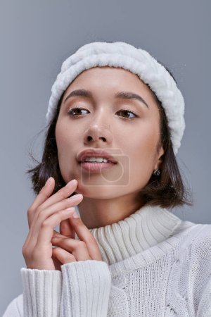 romantische und verträumte asiatische Frau im Winterpullover und warmweißem Stirnband, die wegschaut auf grau
