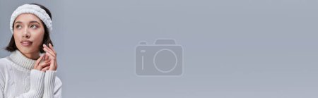 Téléchargez les photos : Modèle asiatique à la mode et rêveuse en pull blanc et bandeau chaud regardant loin sur gris, bannière - en image libre de droit