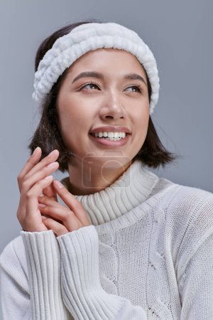 Porträt einer unbeschwerten Asiatin in Strickpullover-Stirnband, die vom grauen, glücklichen Winter wegblickt