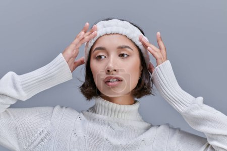stylish asian woman in knitted sweater adjusting warm headband on grey, winter fashion photography