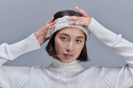 young asian woman in white sweater adjusting soft headband on grey, winter fashion concept