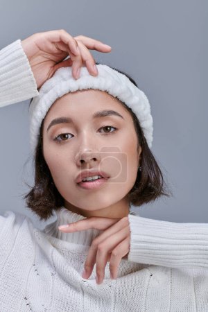 portrait of asian woman with snowy hair posing in white winter sweater and warm headband on grey