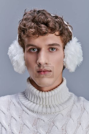 Porträt eines jungen Mannes mit Schnee auf welligem Haar, der in warmen Ohrenschützern und weißem Pullover auf grau posiert