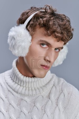 confident man in winter earmuffs and white knitted sweater looking at camera on grey, seasonal style