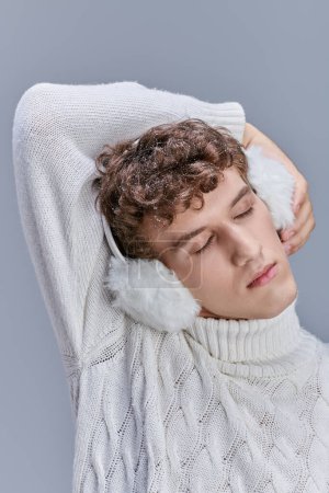 junger Mann in weißem Strickpullover und Ohrenschützer posiert mit geschlossenen Augen auf grauer, gemütlicher Wintermode