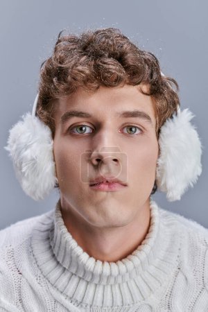Mann mit welligem, schneebedecktem Haar und ausdrucksstarkem Blick trägt Winterohrenschützer und blickt in die Kamera auf grau