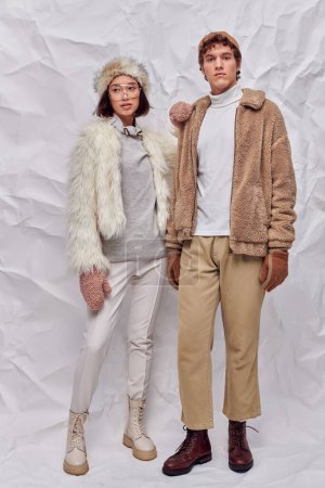 longitud completa de la pareja multiétnica en ropa acogedora posando sobre fondo de textura blanca, moda de invierno