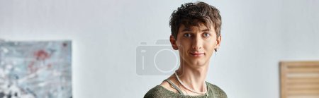 happy transgender person in earring and pearl necklace looking at camera in modern apartment, banner