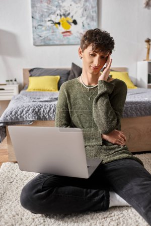 curly transgender influencer using laptop and sitting on carpet near bed, blogger using social media