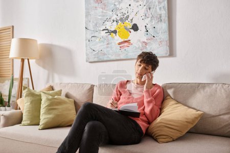 Transgender-Bloggerin im rosafarbenen Pullover, die auf dem Sofa sitzt und mit dem Smartphone spricht und in ein Notizbuch schreibt