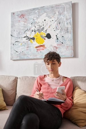 blogueur transgenre en pull rose tenant smartphone et écriture dans un cahier, assis sur le canapé