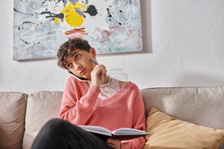 transgender person in pink sweater talking on smartphone and writing in notebook, sitting on sofa
