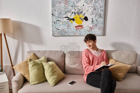 glückliche Transgender-Bloggerin im rosafarbenen Pullover, die in Notizbuch schreibt und auf dem Sofa aufs Smartphone schaut