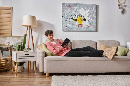 happy transgender person in pink sweater writing in notebook and sitting on sofa next to smartphone