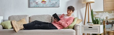 happy transgender influencer in pink sweater writing in notebook and sitting on sofa, banner