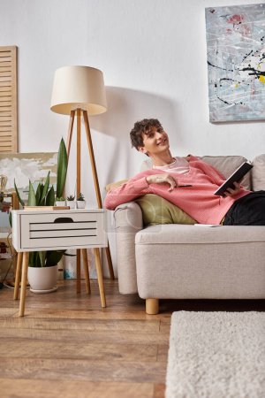 happy transgender person in pink sweater holding notebook and sitting on sofa next to smartphone