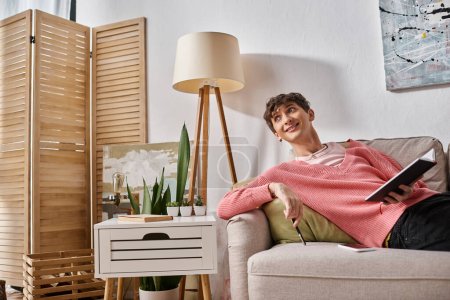 cheerful transgender person in pink sweater holding notebook and sitting on sofa next to smartphone