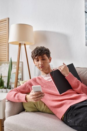 curly transgender person in pink sweater holding notebook, using smartphone and sitting on sofa