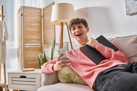 amazed transgender person in pink sweater holding notebook, using smartphone and sitting on sofa