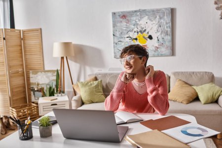 happy transgender freelancer in pink sweater looking away near laptop on desk, remote work