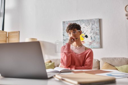 junge Transgender-Freelancer im rosafarbenen Pullover mit Stift und Blick auf Laptop auf dem Schreibtisch, Fernarbeit