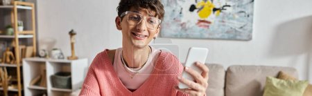 happy androgynous blogger in pink sweater and eyeglasses using smartphone at home, horizontal banner