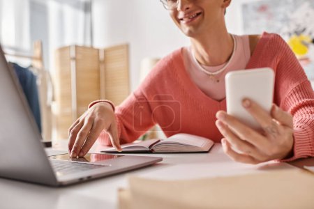 cropped androgynous freelancer in pink sweater and eyeglasses holding smartphone and using laptop