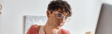 curly androgynous freelancer in pink sweater and eyeglasses looking at laptop, horizontal banner