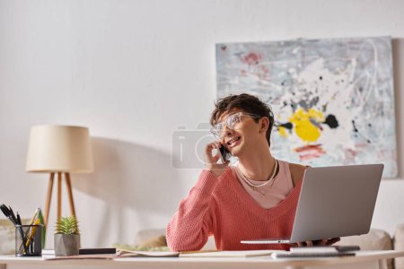 pleased androgynous freelancer in pink sweater and eyeglasses using laptop and smartphone at home