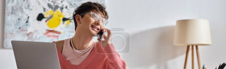 cheerful androgynous freelancer in pink sweater and eyeglasses using laptop and smartphone, banner